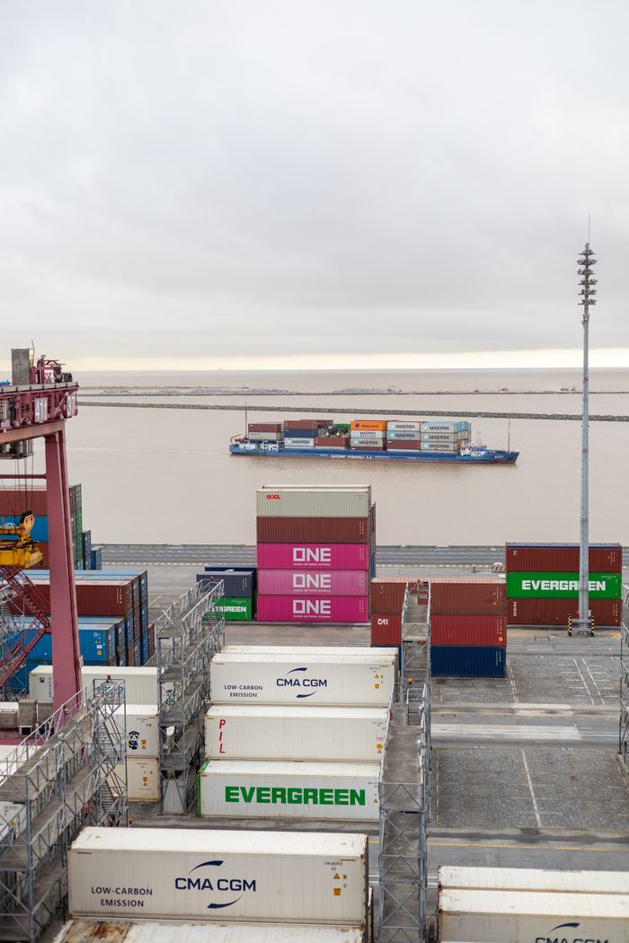 Containers stacked at a port with a cargo ship passing by on a cloudy day.